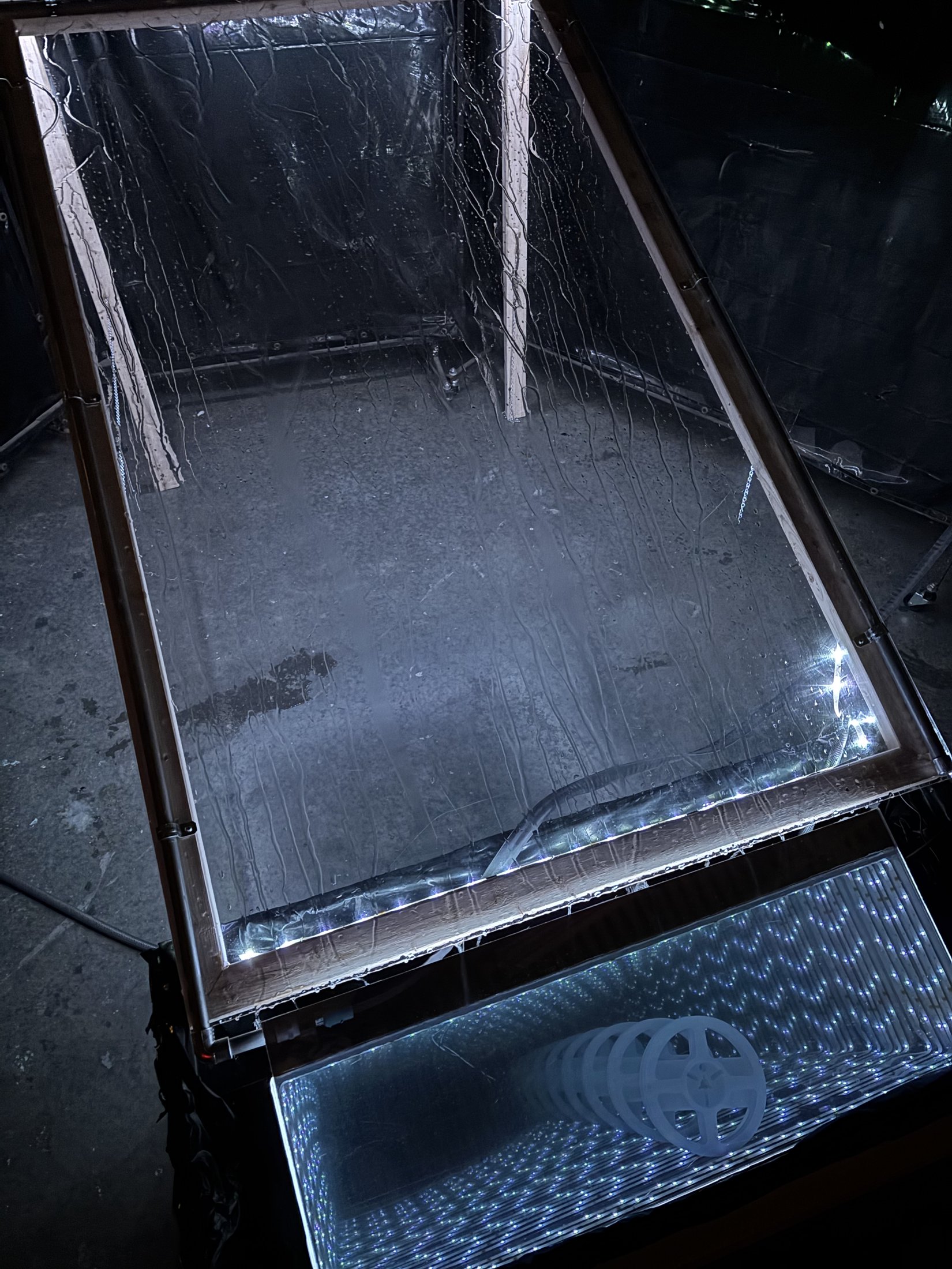 The sculpture seen from above. A wet sheet of plexiglass in a wooden frame slopes down to an infinity mirror, where rows of white lights recede into apparent depth.