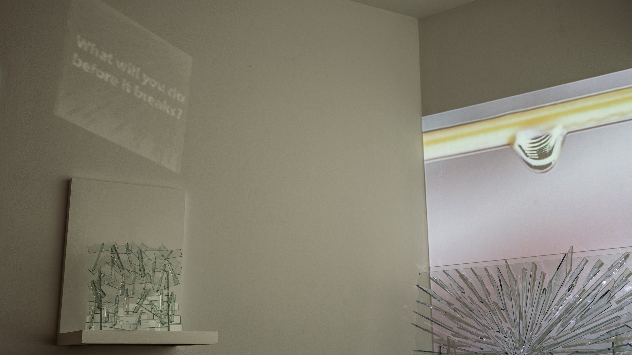 A small sculpture of stacked glass shards on a white shelf. Above it, a reflected projection reads What will you do before it breaks? To the right, a projected water droplet above the radial glass sculpture.