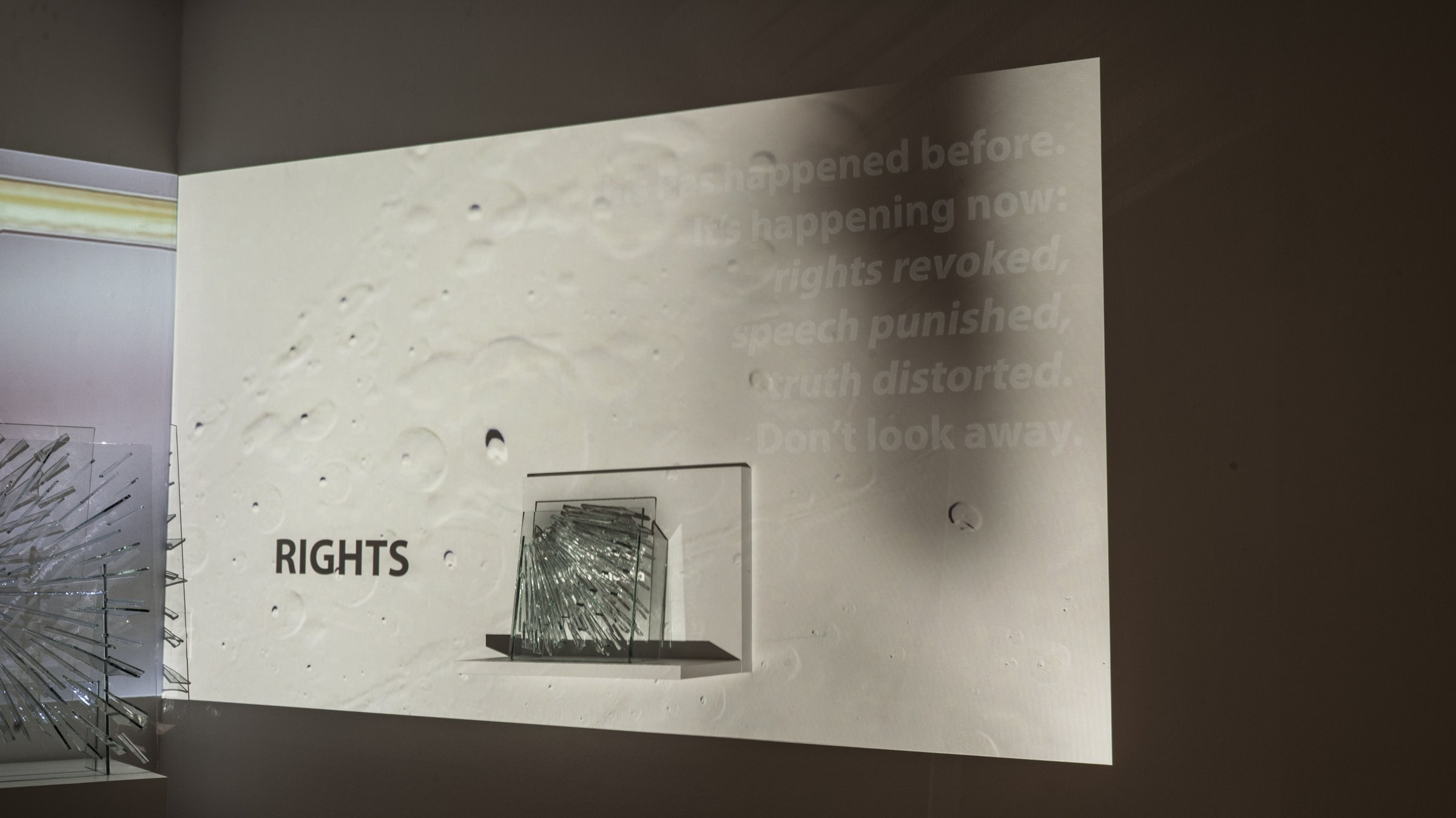 A glass sculpture on a shelf inside a projection, beside the projected word RIGHTS. A shadow falls across the wall and faint text shows inside it.